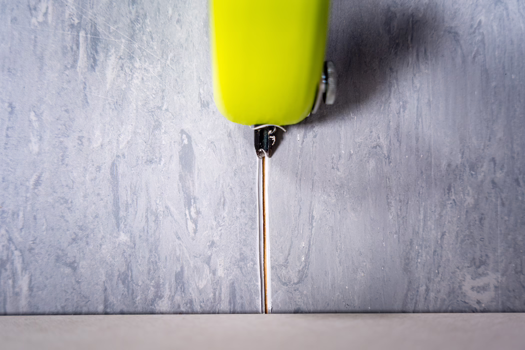 Gros plan d'un cutter rétractable vert citron positionné verticalement au-dessus d'un joint étroit entre un mur gris texturé et un sol clair. La fine lame métallique est déployée vers le bas dans le joint, avec le manche en plastique et une petite vis métallique visibles en haut du cadre. La scène met en valeur les textures contrastées — plâtre peint rugueux du mur et surface lisse du sol — et est éclairée doucement depuis le dessus, révélant de légères rayures et traces d'usure sur la lame et le manche.