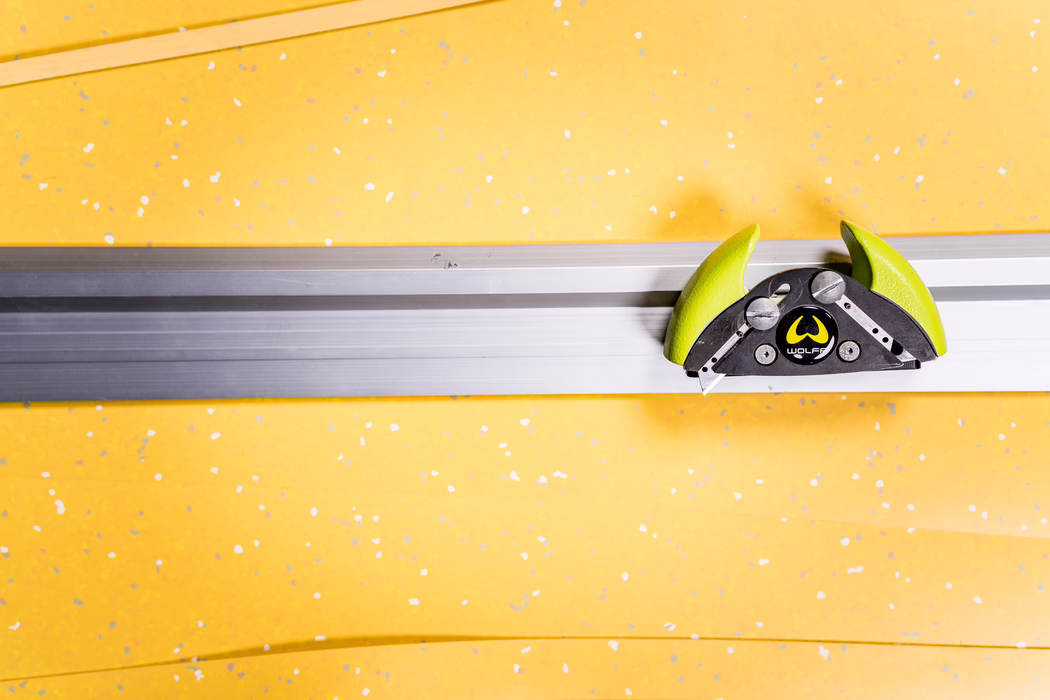 WOLFF_Application Image_Railcut_Detail2_2022-11 Gros plan d'un rail horizontal en aluminium fixé sur un mur jaune vif parsemé de taches blanches et grises. Sur la droite, une pince coulissante verte et noire de marque Wolff est fixée au rail ; ses poignées lime arrondies, son corps métallique noir, ses vis apparentes et une petite lame rétractable sont bien visibles. La composition met en valeur des lignes horizontales prononcées, des couleurs contrastées et une esthétique industrielle minimaliste.
